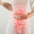 A imagem apresenta uma pessoa vestindo uma camiseta branca com as mãos posicionadas sobre o abdômen. Sobre a região abdominal, há uma ilustração translúcida do trato gastrointestinal, destacando o intestino grosso em uma tonalidade vermelha, sugerindo a presença de desconforto ou problema intestinal.