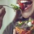 A imagem mostra uma pessoa prestes a dar uma mordida em uma salada colorida. Ela está segurando uma tigela de vidro cheia de vegetais frescos, incluindo alface, tomate, cenoura ralada e pedaços de queijo. A pessoa está usando brincos grandes e aparenta estar saboreando a refeição saudável com um sorriso.