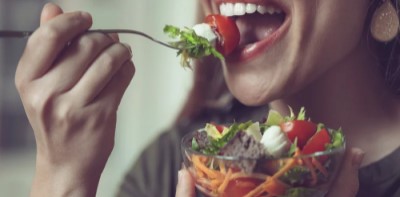A imagem mostra uma pessoa prestes a dar uma mordida em uma salada colorida. Ela está segurando uma tigela de vidro cheia de vegetais frescos, incluindo alface, tomate, cenoura ralada e pedaços de queijo. A pessoa está usando brincos grandes e aparenta estar saboreando a refeição saudável com um sorriso.