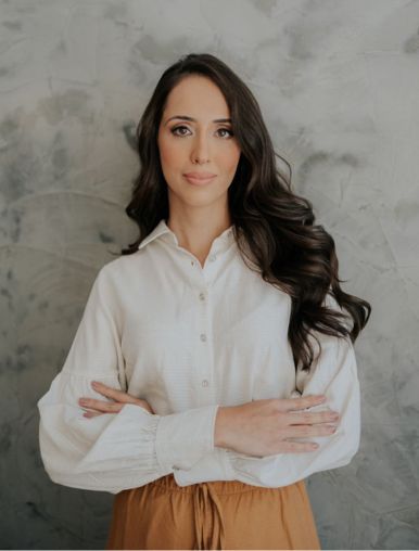 A imagem mostra Dra Thais sorridente com cabelos castanhos longos e ondulados, vestindo uma blusa preta. O fundo é de uma cor neutra, destacando seu rosto e sua expressão amigável e confiante. Dra Thais está olhando diretamente para a câmera, transmitindo uma sensação de profissionalismo e acolhimento.