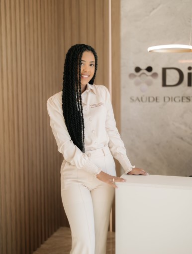 Na imagem, uma mulher sorridente com longas tranças está em pé ao lado de um balcão branco. Ela veste uma camisa social clara e calça combinando, transmitindo elegância e profissionalismo. Ao fundo, uma parede com o logotipo de uma clínica de saúde, que sugere um ambiente corporativo ou médico. A iluminação suave e o design moderno do espaço reforçam a sensação de acolhimento e sofisticação.