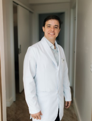 Na imagem, Doutor César sorri enquanto veste um jaleco branco, típico de profissionais da área da saúde. Ele está em um corredor iluminado, transmitindo uma imagem de profissionalismo e cordialidade. Com cabelo curto e escuro, ele posa de forma confiante, sugerindo que está pronto para atender ou realizar um procedimento clínico. A expressão no rosto transmite simpatia e acolhimento.