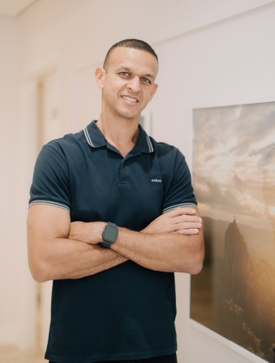 Na imagem, um homem sorri com os braços cruzados, vestindo uma camisa polo azul marinho e um relógio esportivo no pulso. Ele está em pé em um ambiente bem iluminado, ao lado de uma parede com uma obra de arte que retrata uma paisagem. Sua expressão é descontraída e confiante, transmitindo simpatia e acolhimento.