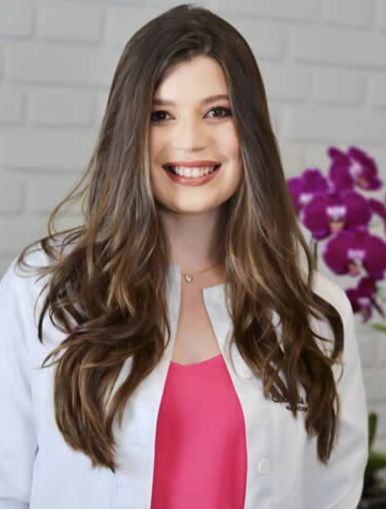 A imagem mostra Gabriela que tem cabelos longos, usa um jaleco branco e blusa rosa. Ela sorri e olha diretamente para câmara, transmitindo profissionalismo e confiança.