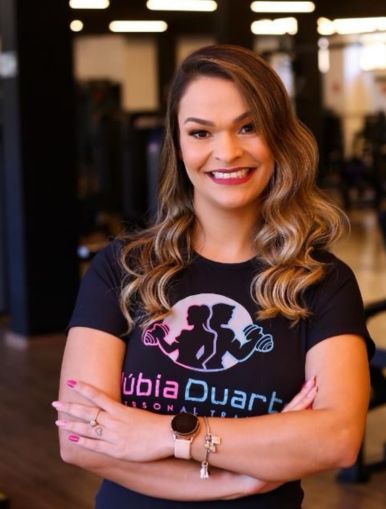 Uma mulher sorridente posa em uma academia, cruzando os braços com confiança. Ela tem cabelos longos e ondulados em tom loiro com raiz escura e usa uma camiseta preta com o logo "Núbia Duarte Personal Trainer". Suas unhas estão pintadas de rosa, e ela usa um relógio no pulso esquerdo. O ambiente ao fundo é bem iluminado, com equipamentos de musculação visíveis, reforçando o cenário de treino e bem-estar.